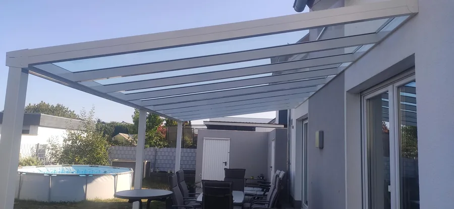 Eine moderne Terrassenüberdachung aus Glas und Metall über einem Essbereich im Freien mit Pool im Hintergrund.